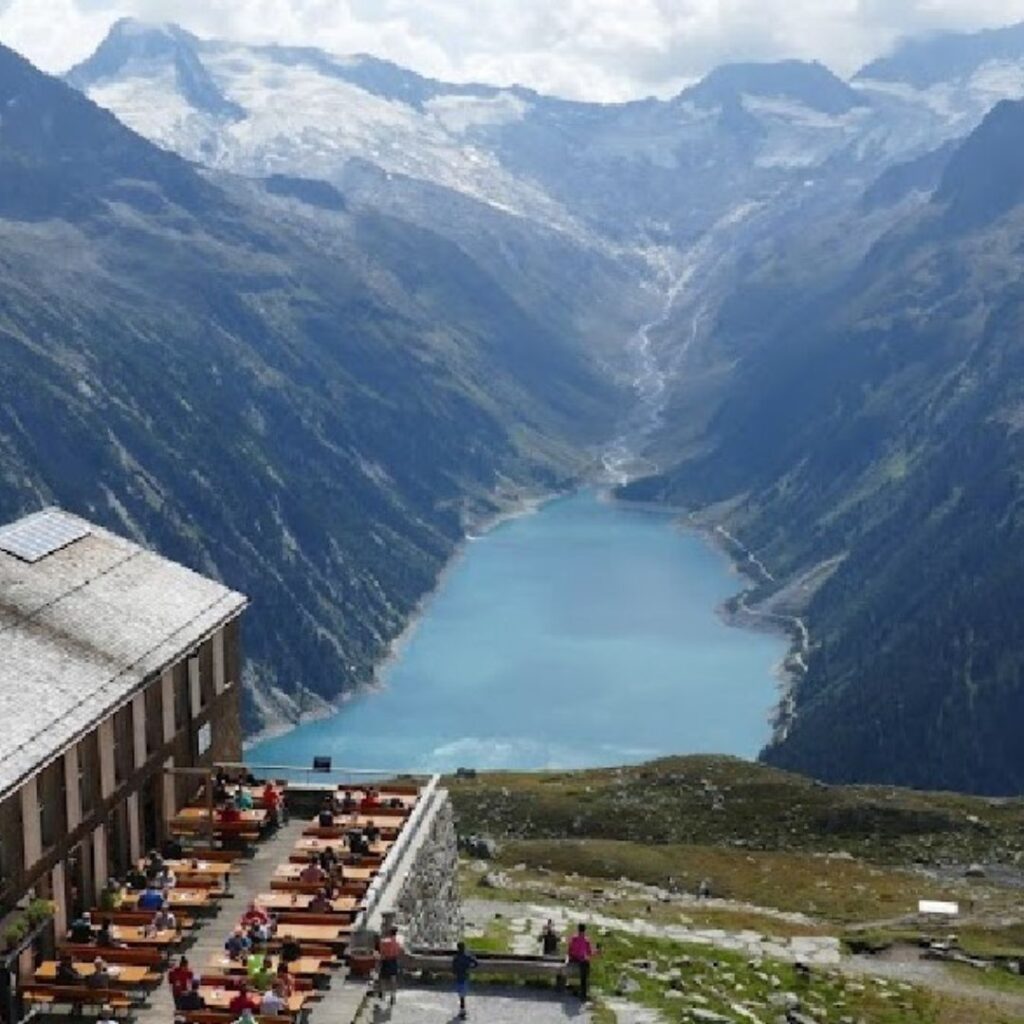 Panoramablick auf den Schlegeis Stausee, eingebettet zwischen hohen, schneebedeckten Alpenbergen, mit einem Berghaus im Vordergrund.