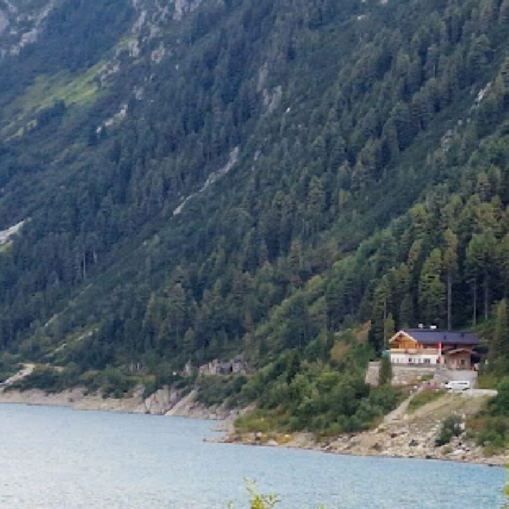 Ein Haus am Ufer des Schlegeis Stausees, eingebettet in eine bewaldete Berglandschaft mit steilen Berghängen.