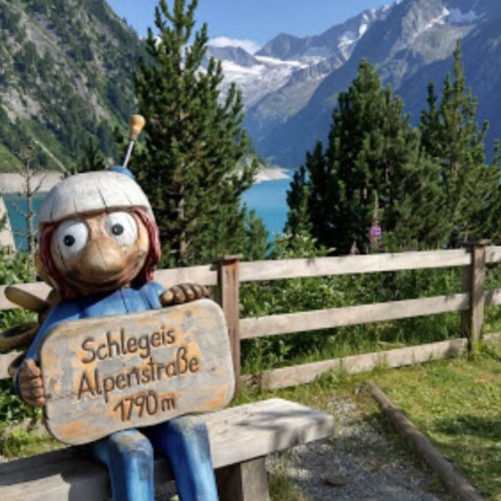 Schlegeis Alpenstraße mit einem Holzmaskottchen, das ein Schild mit der Aufschrift "Schlegeis Alpenstraße 1790m" hält. Im Hintergrund sind Berge und der Stausee zu sehen.