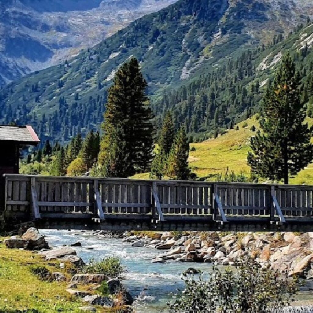 Holzbrücke, die einen klaren Gebirgsbach inmitten einer alpinen Landschaft überquert, umgeben von hohen Bäumen und Bergen.