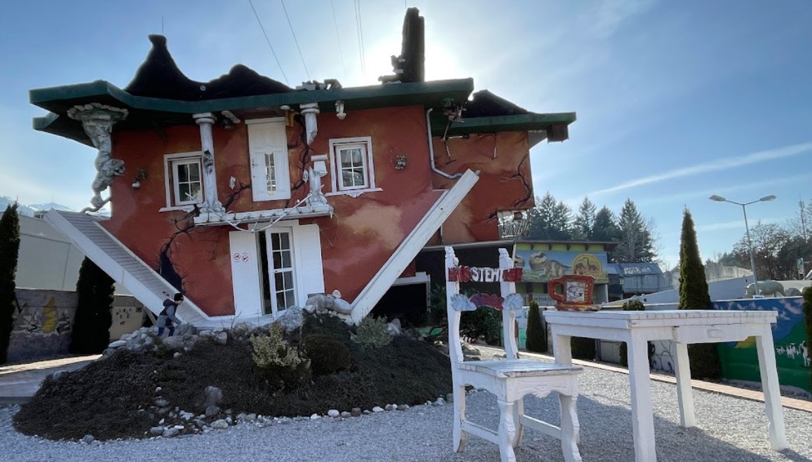 Umgedrehtes Haus mit skurrilen, schiefen Elementen und einem großen Tisch im Vordergrund bei strahlend blauem Himmel